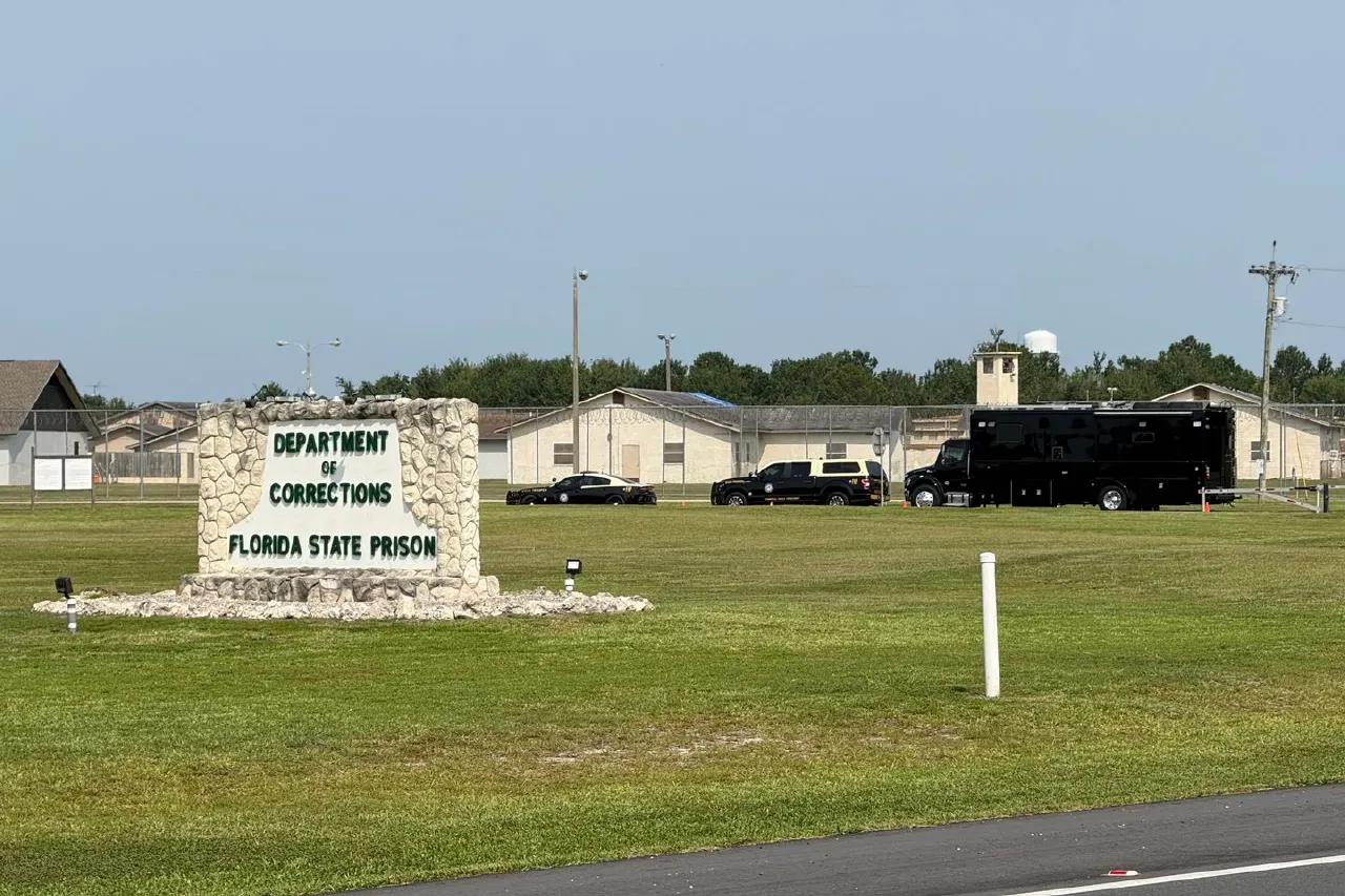 Entrance sign for Florida State Prison with Department of Corrections, prison buildings and vehicles behind.