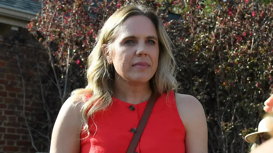 Woman in red sleeveless top with brown strap outdoors, brick wall and shrubs behind her.