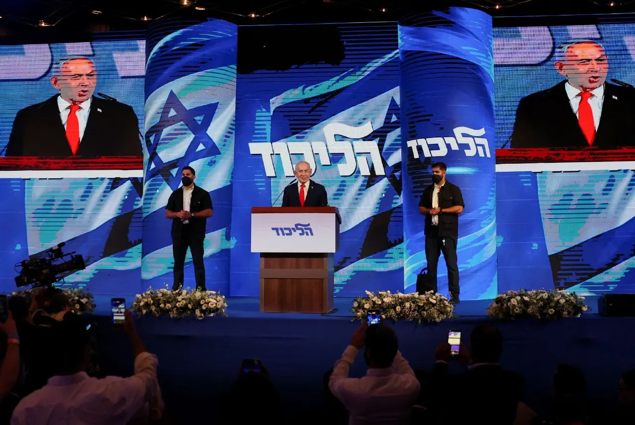 Man speaking at Likud podium before large blue Likud banners, Israeli flags and screens, with audience filming.