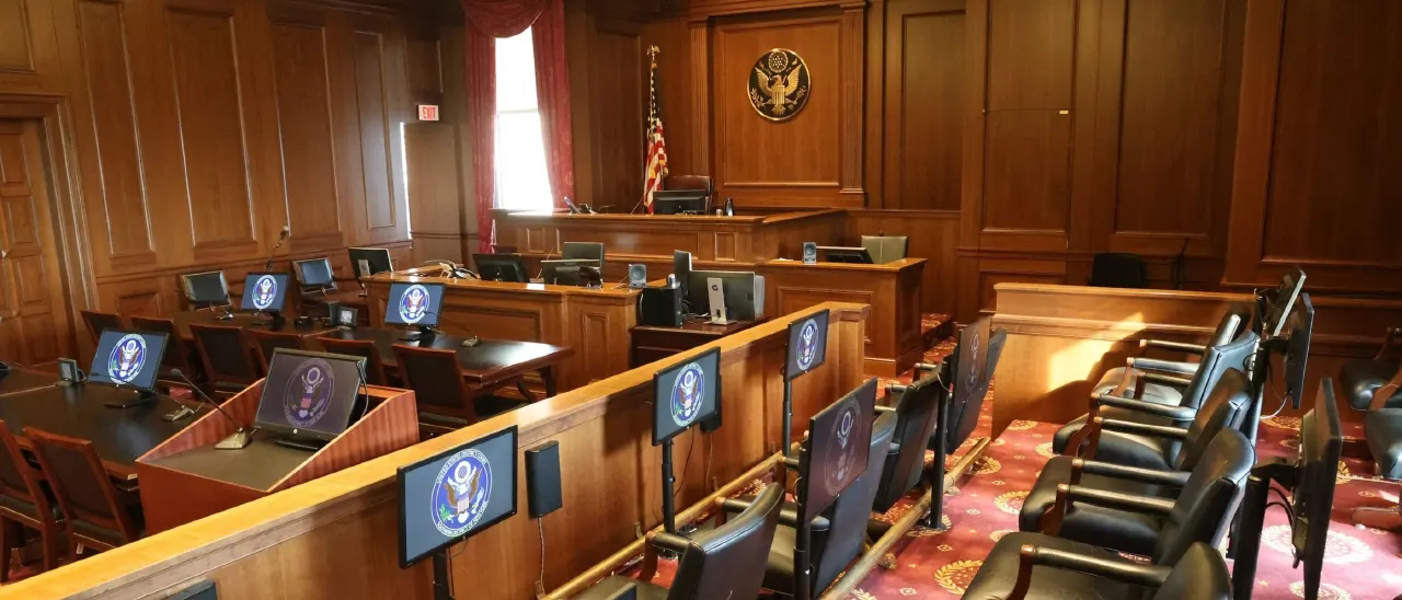 Empty wood-paneled courtroom with judge's bench, monitors, flag, and seal emblem.