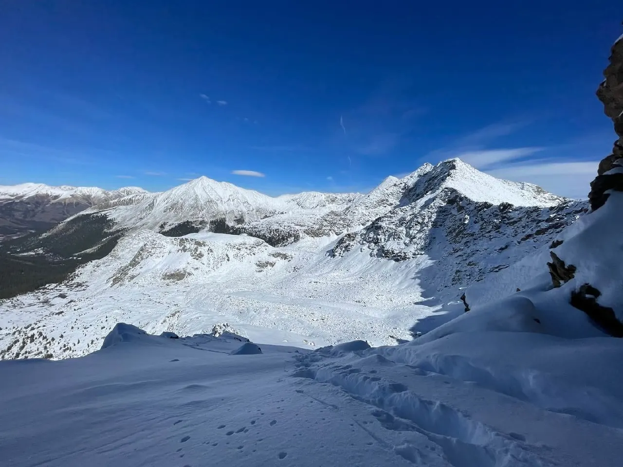 Snow-covered mountain ridges under a bright blue sky, with footprints in fresh snow.