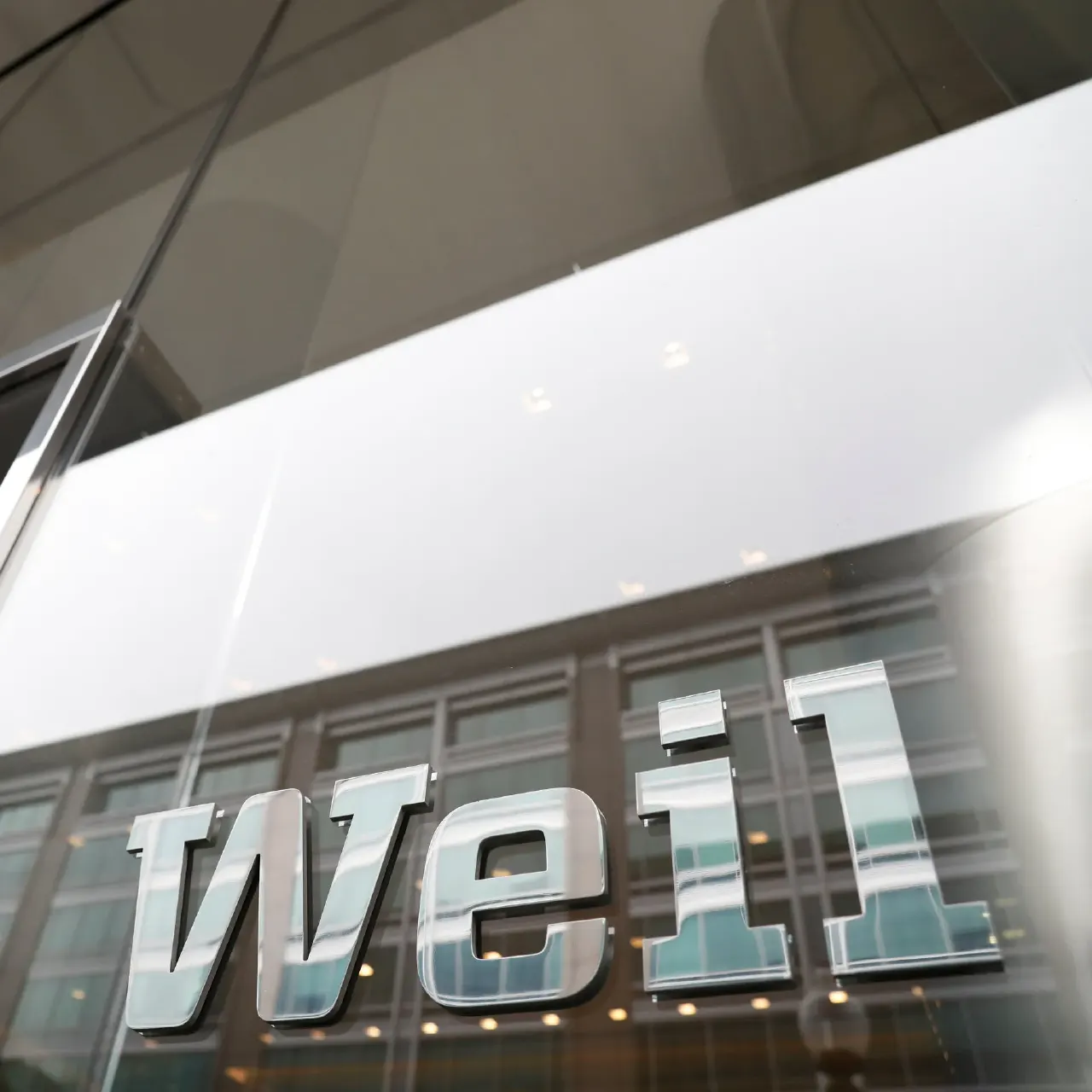 Metal 'weil' lettering mounted on a glass office building facade, viewed from below.