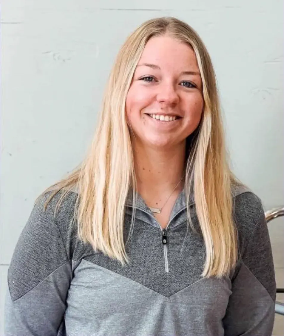 Smiling young woman with long blonde hair in grey quarter-zip top against pale wall.