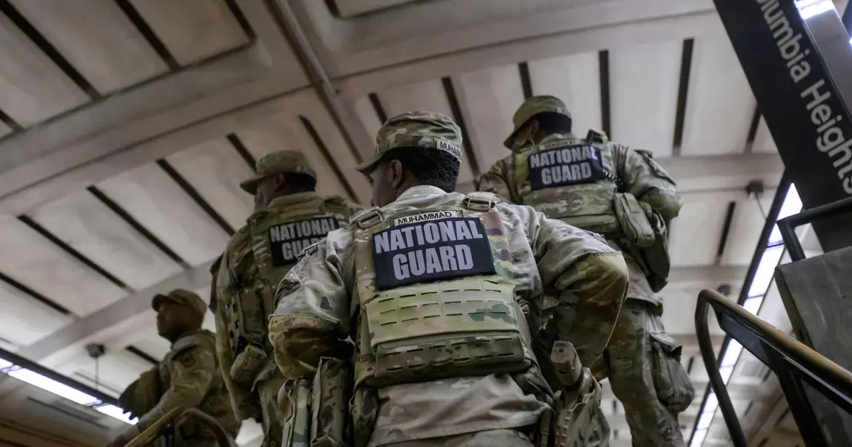 Uniformed National Guard members in camouflage with reflective vests walking up a station staircase, backs to camera.