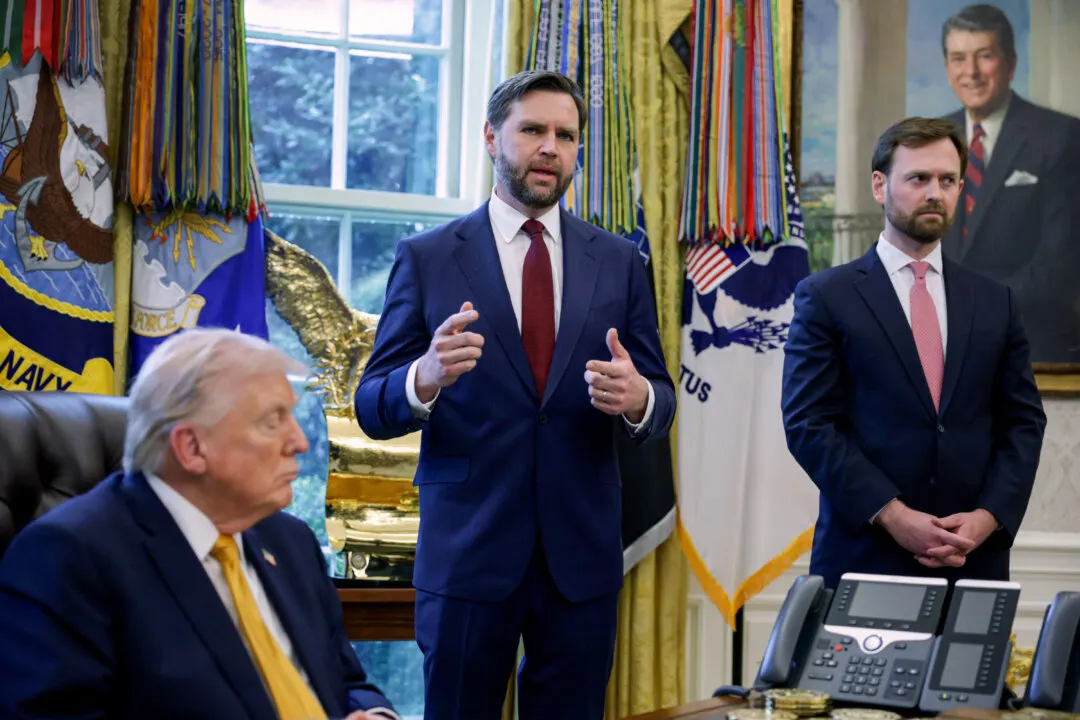 Men in suits in an ornate office; one speaks, one seated, flags and portrait behind.