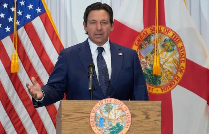 Man in dark suit speaking at podium with Florida state seal, beside US and Florida flags.