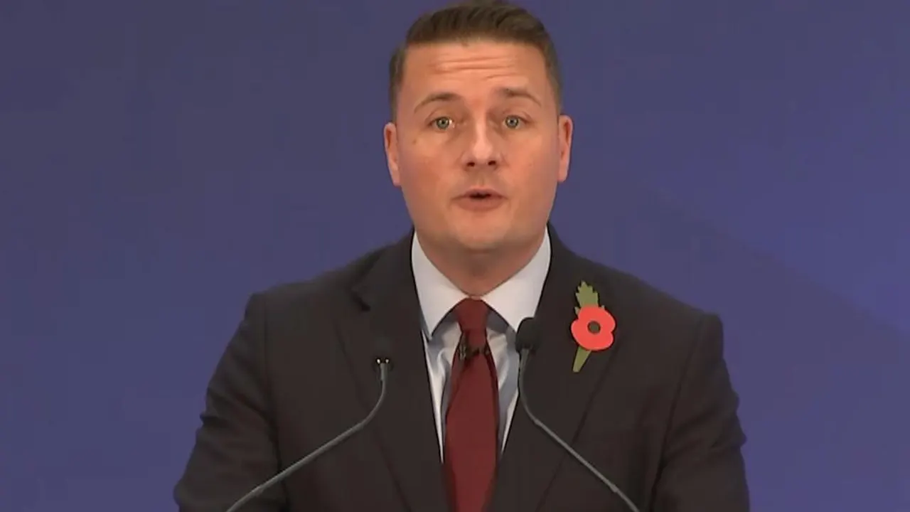 A man in a dark suit and red tie with a poppy speaks at a podium before a blue backdrop.