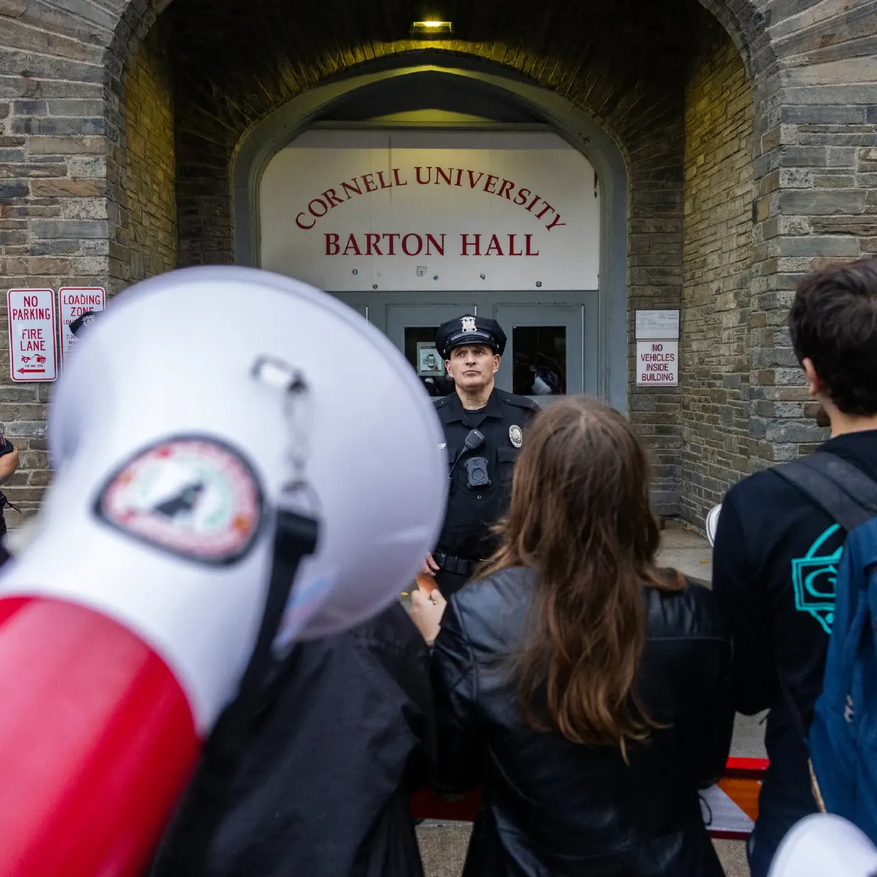 Police officer stands at Cornell University Barton Hall entrance as people with a megaphone confront him.