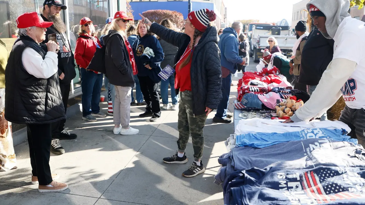 Crowd of people on a street near merchandise tables selling red caps, flag-print shirts and apparel.