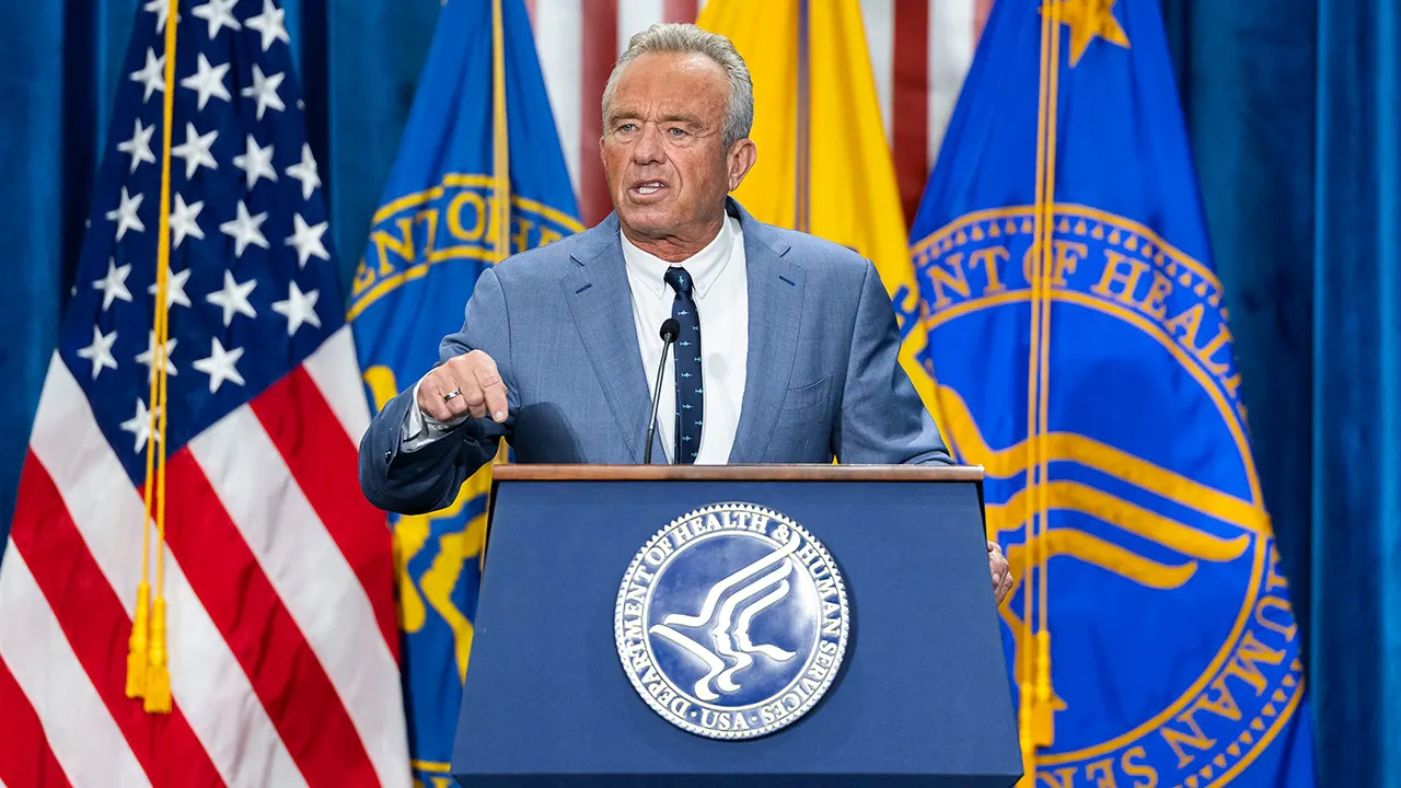 Man in gray suit speaking at podium with HHS seal, flags behind him