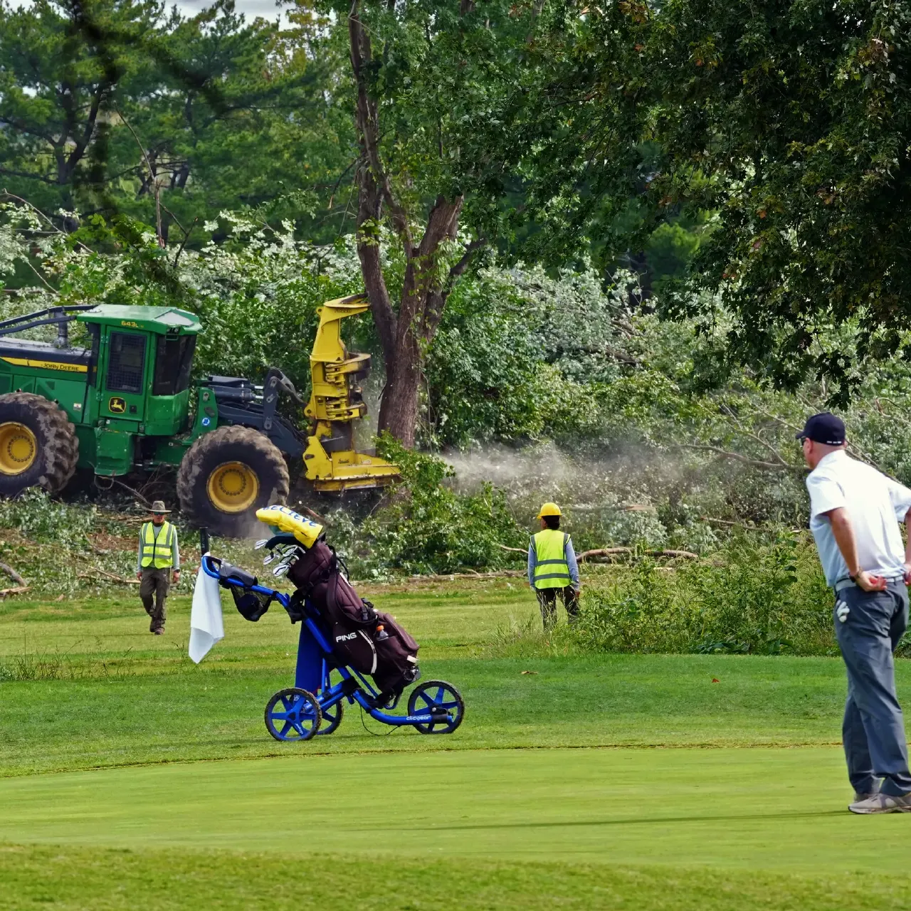 Golf course with tree-clearing machinery, workers in hi-vis vests, golf cart and golfer.
