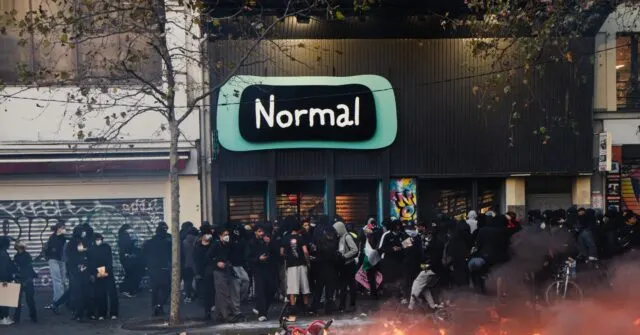 Crowd of masked, hooded people on a street in front of a shopfront with red smoke.