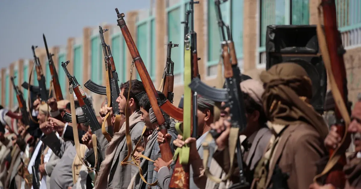 Row of armed men in traditional dress raising rifles outdoors in front of a building.