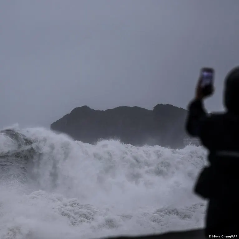 Large waves crash against a dark rocky coastline as a silhouetted person films with a phone.
