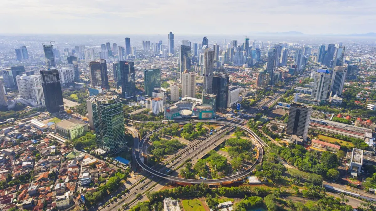 Aerial view of Jakarta skyline with high-rise towers, a roundabout expressway and dense urban neighborhoods.