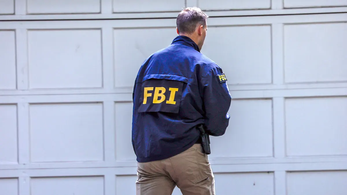 Man in navy FBI jacket with holstered weapon, facing away from camera toward a garage door.