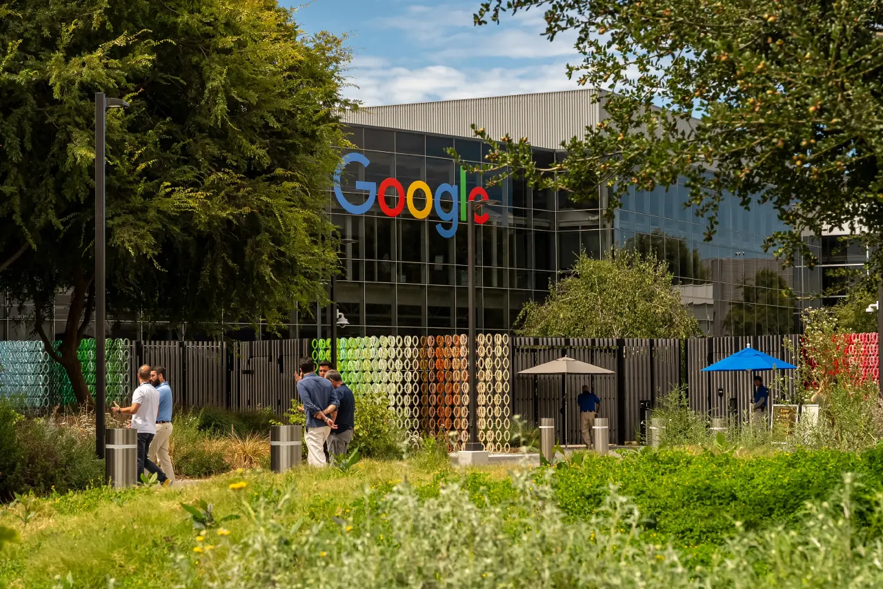 Google logo on glass office building with people chatting outside amid greenery and umbrellas.