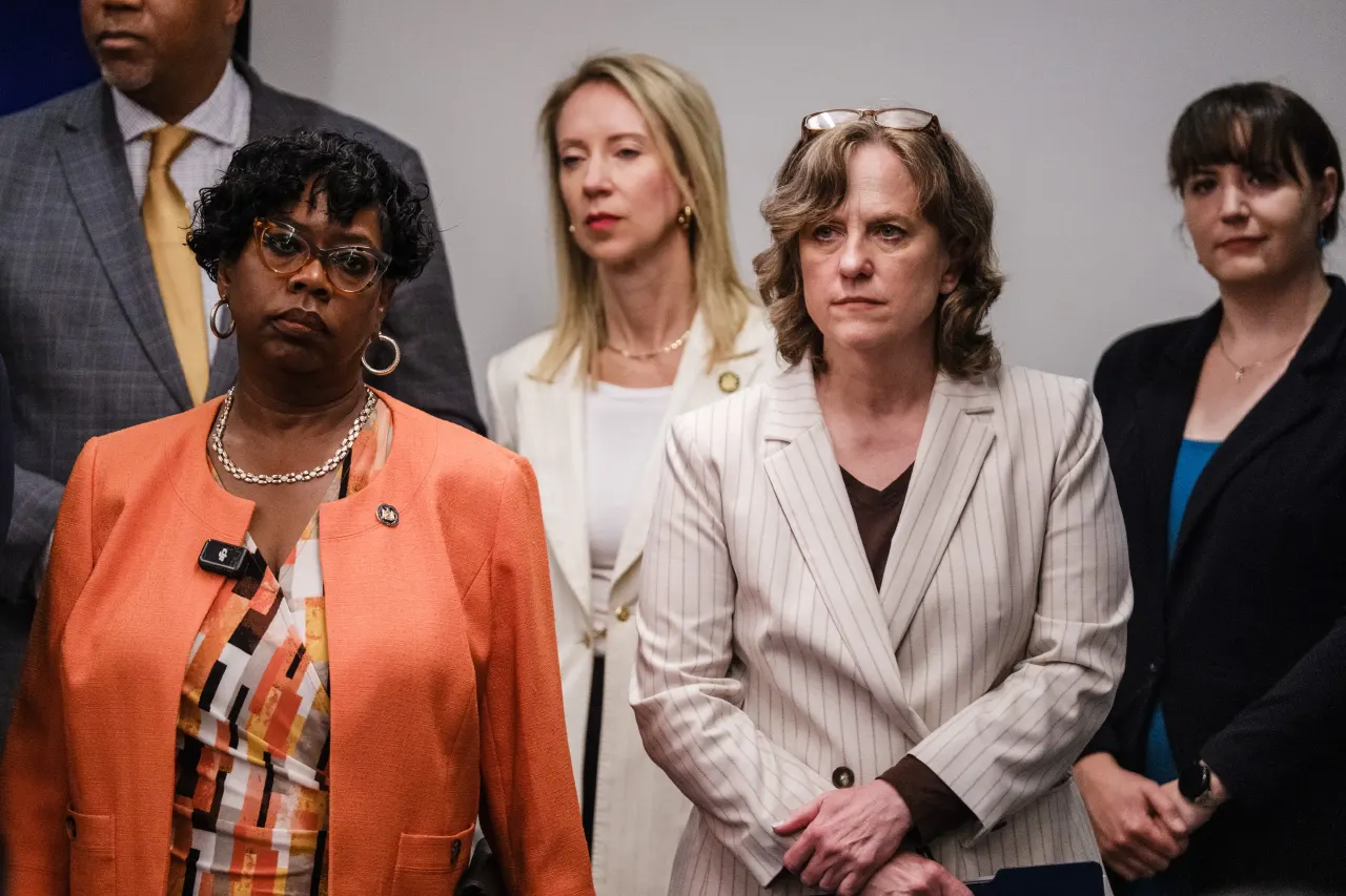 Four women and a man stand solemnly indoors at what appears to be a press event.