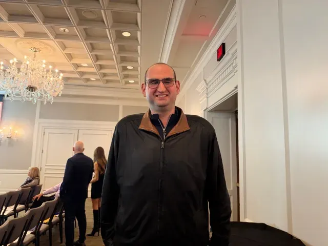 Smiling man in glasses and black jacket standing in a formal event room with chandelier.