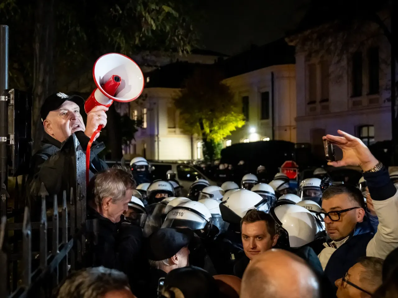 Crowd at night with a man using a megaphone facing rows of helmeted police behind a fence.