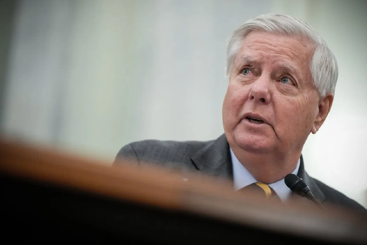 Older man with white hair in gray suit speaking at a microphone, shot from below.