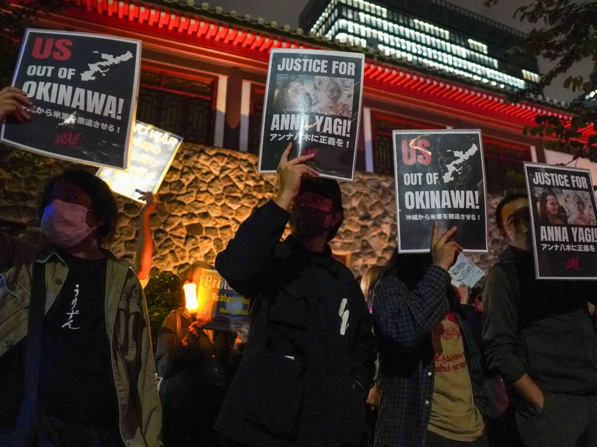 Night protest in Okinawa, demonstrators holding 'US out of Okinawa' and 'Justice for Anna Yagi' placards.