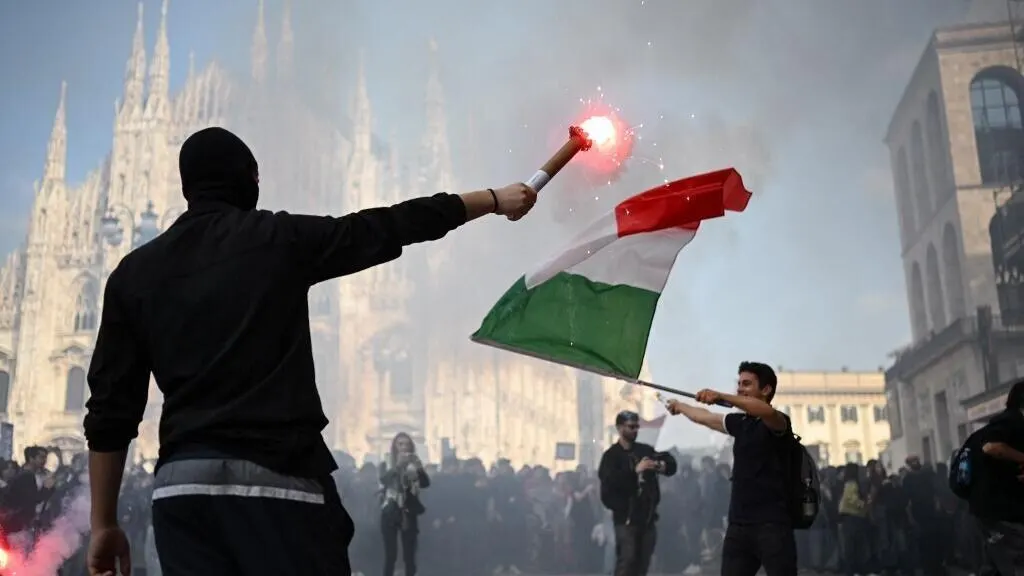 Masked protester holds flare beside Italian flag before Milan cathedral amid crowd and smoke.