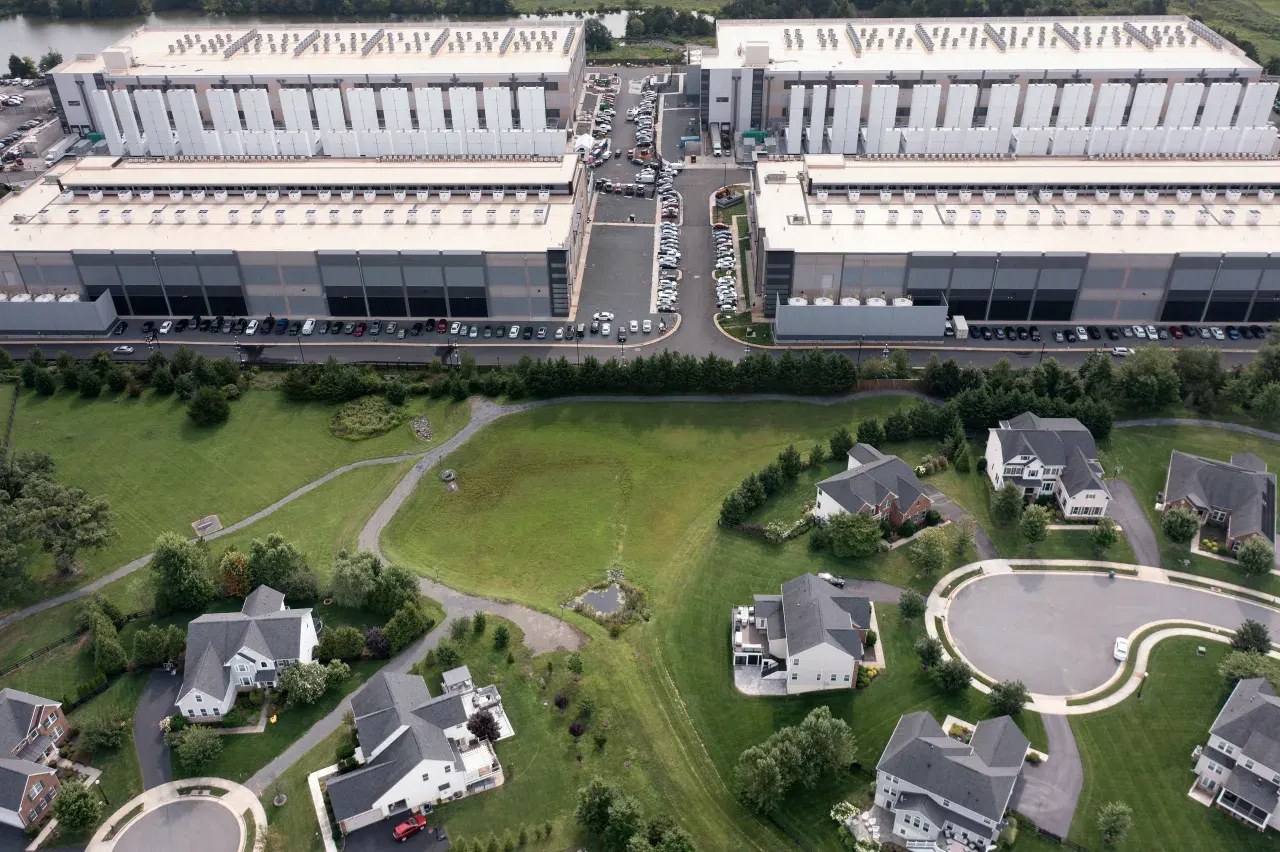 Aerial view of large data center warehouses beside a residential neighborhood with houses and parking lots.