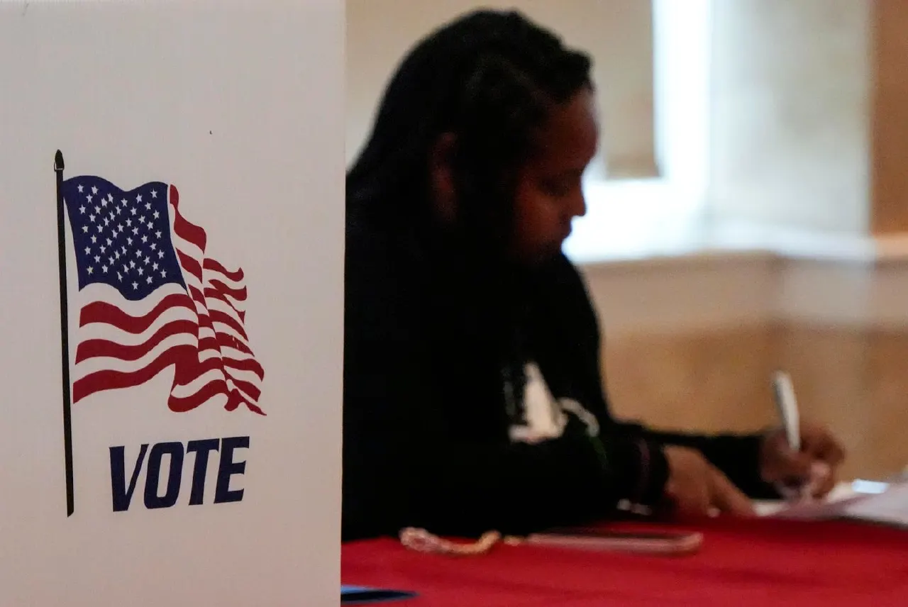 Voter writing at a table beside a white VOTE sign with an American flag graphic.
