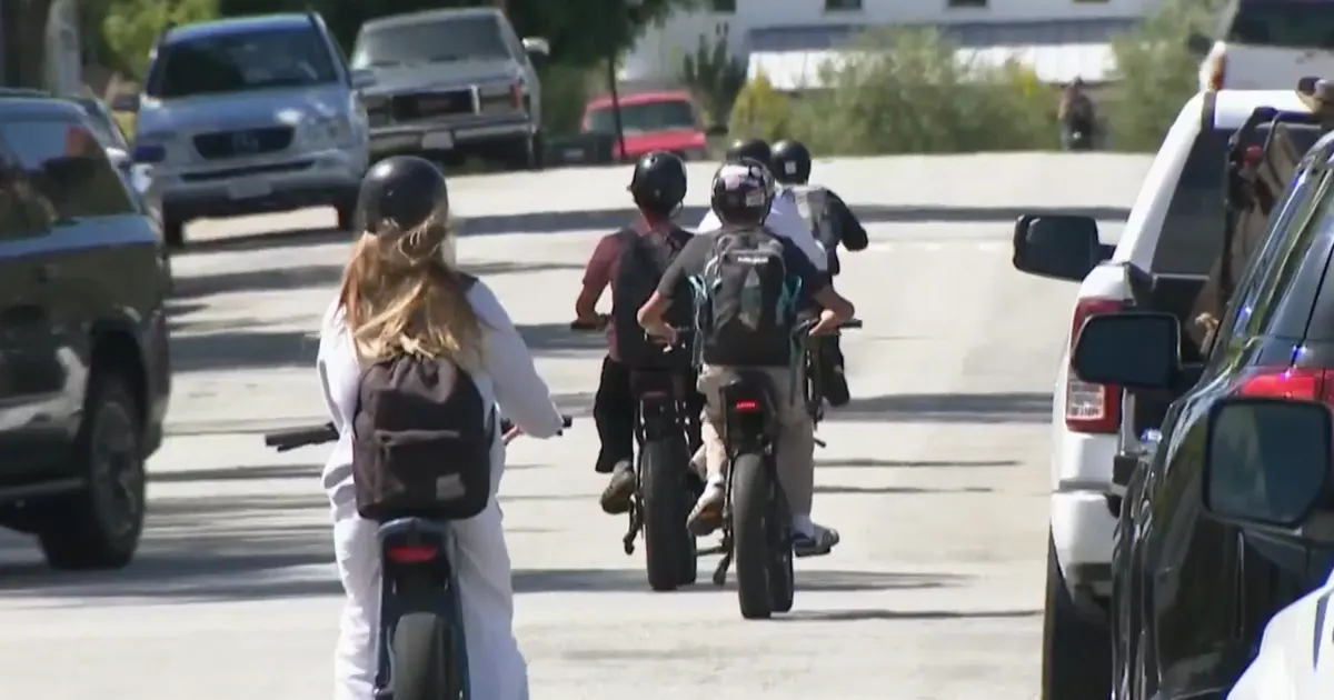 Several people riding electric bikes down a street, shot from behind, with parked cars