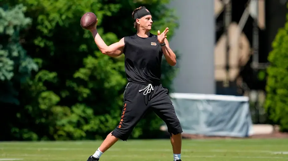 Football player in black Bengals practice gear throwing a football on a grass field.