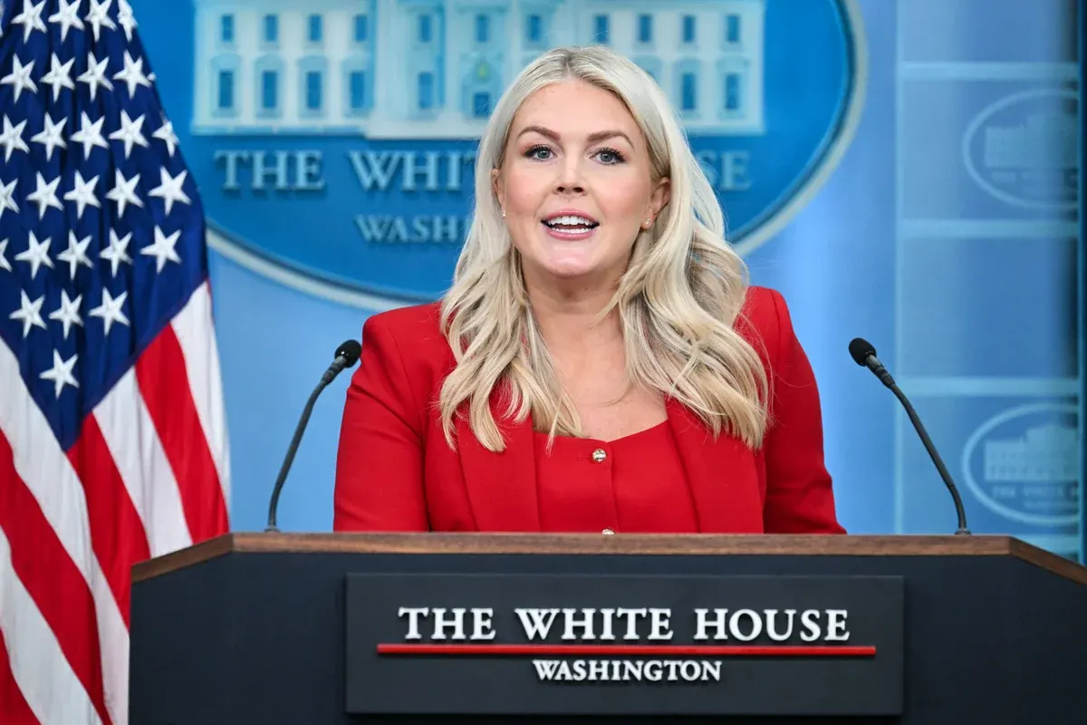 Woman in red blazer speaking at White House press briefing podium before American flag and seal.