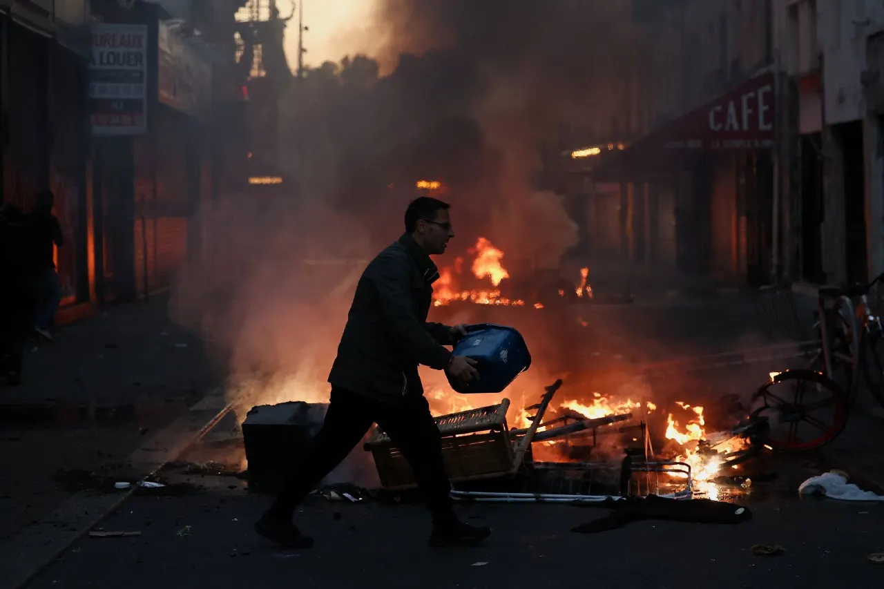 Man carrying a blue bin past burning debris and fire on a smoke-filled city street