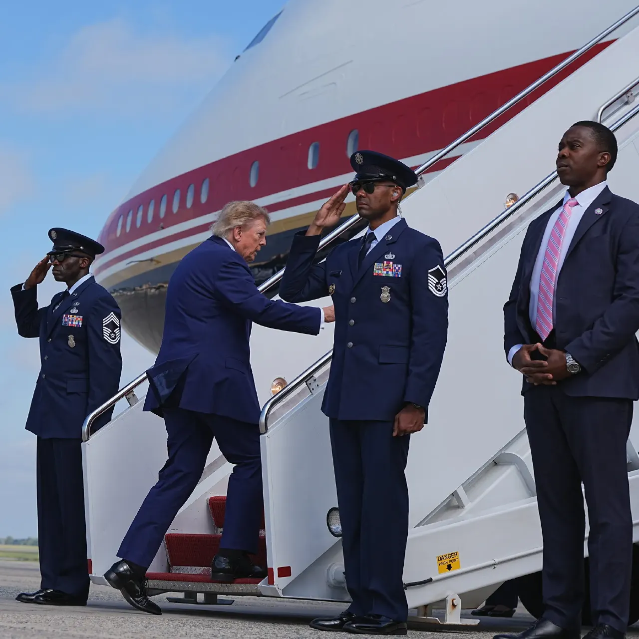 Man in dark suit boarding a red-striped aircraft as uniformed officers salute beside the stairs.