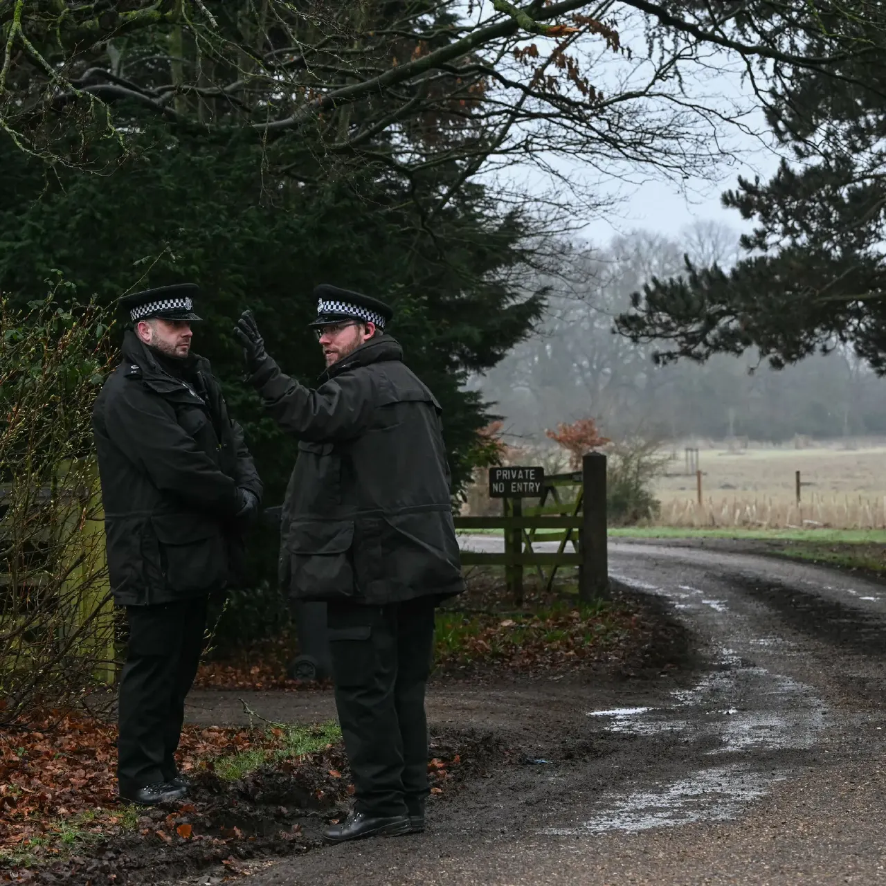 Two uniformed police officers in checked caps talk on a muddy private road by a gate.