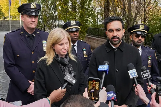 Man speaking to press at microphones beside uniformed police officers, with a blonde woman in foreground.