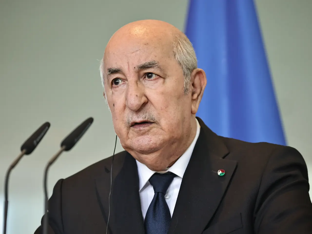 Older man in dark suit speaking at microphones before a blue flag