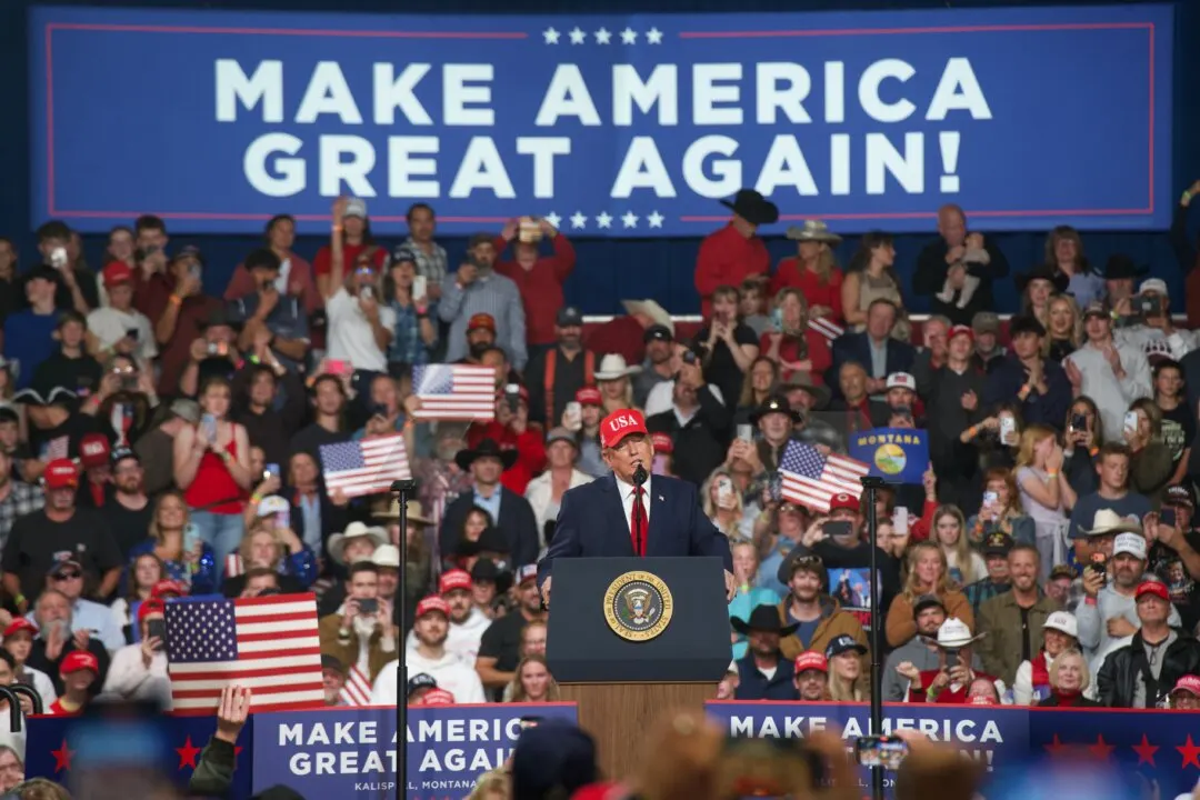 Speaker at a podium with presidential seal before a rally crowd, Make America Great Again banners and flags.