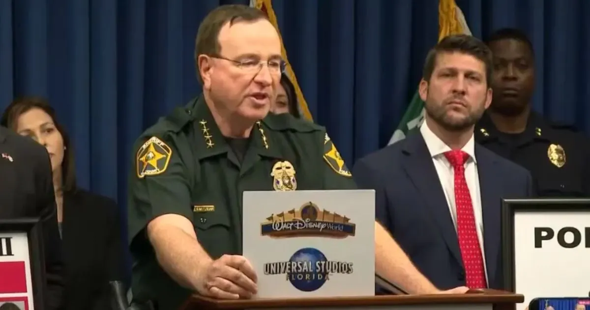 Uniformed sheriff speaking at podium with Disney World and Universal Studios signs, officials behind him.