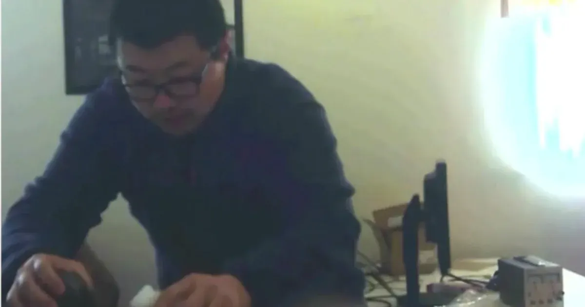 Blurry video still of a man in glasses and dark sweater bent over a desk with monitors.