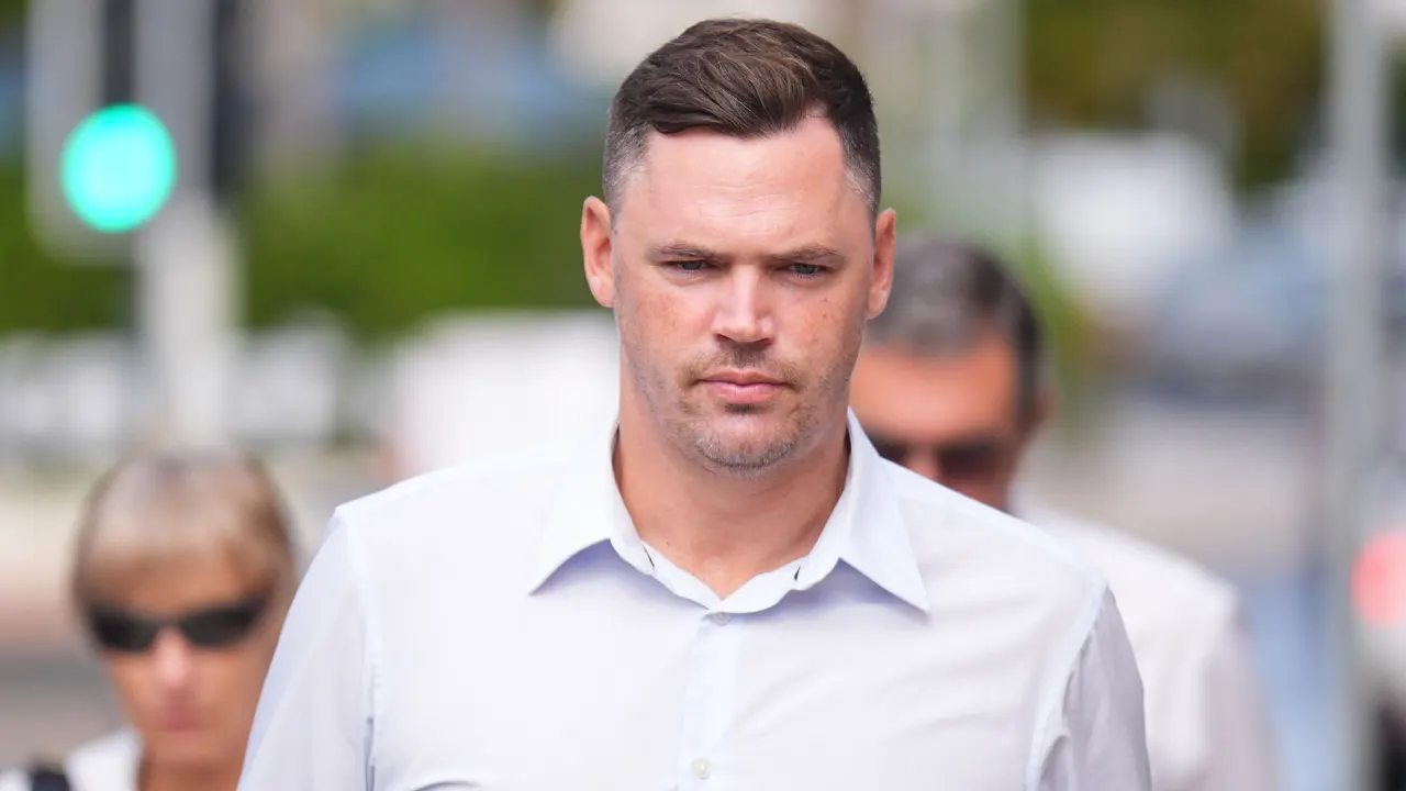 Man in white shirt walking outdoors on a street, with blurred pedestrians and traffic lights behind him.