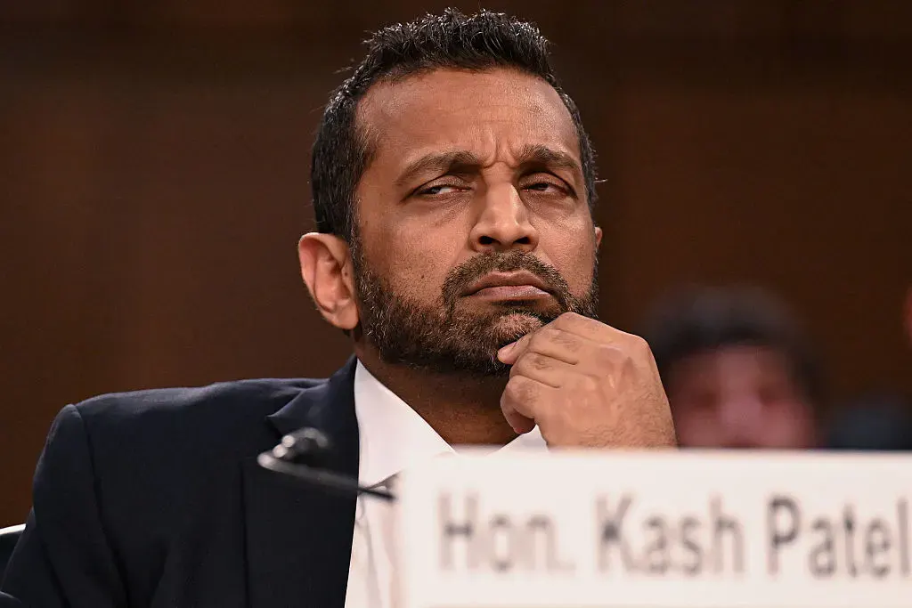 Man in dark suit at hearing table, hand on chin, name placard in front