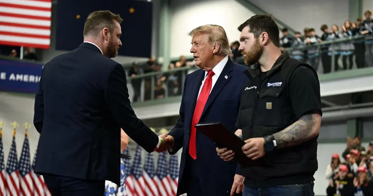 Two men shake hands on stage before American flags, a third man holds a folder, crowd watching.