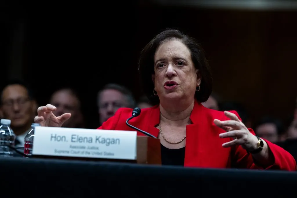 A woman in a red blazer speaks at a hearing table, with a nameplate reading Associate Justice, Supreme Court.