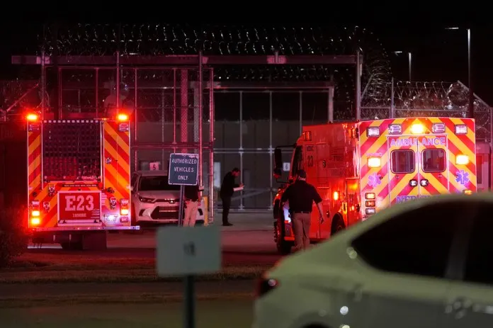 Ambulances and fire vehicle with lights flashing at night outside a prison's fenced, barbed-wire entrance.