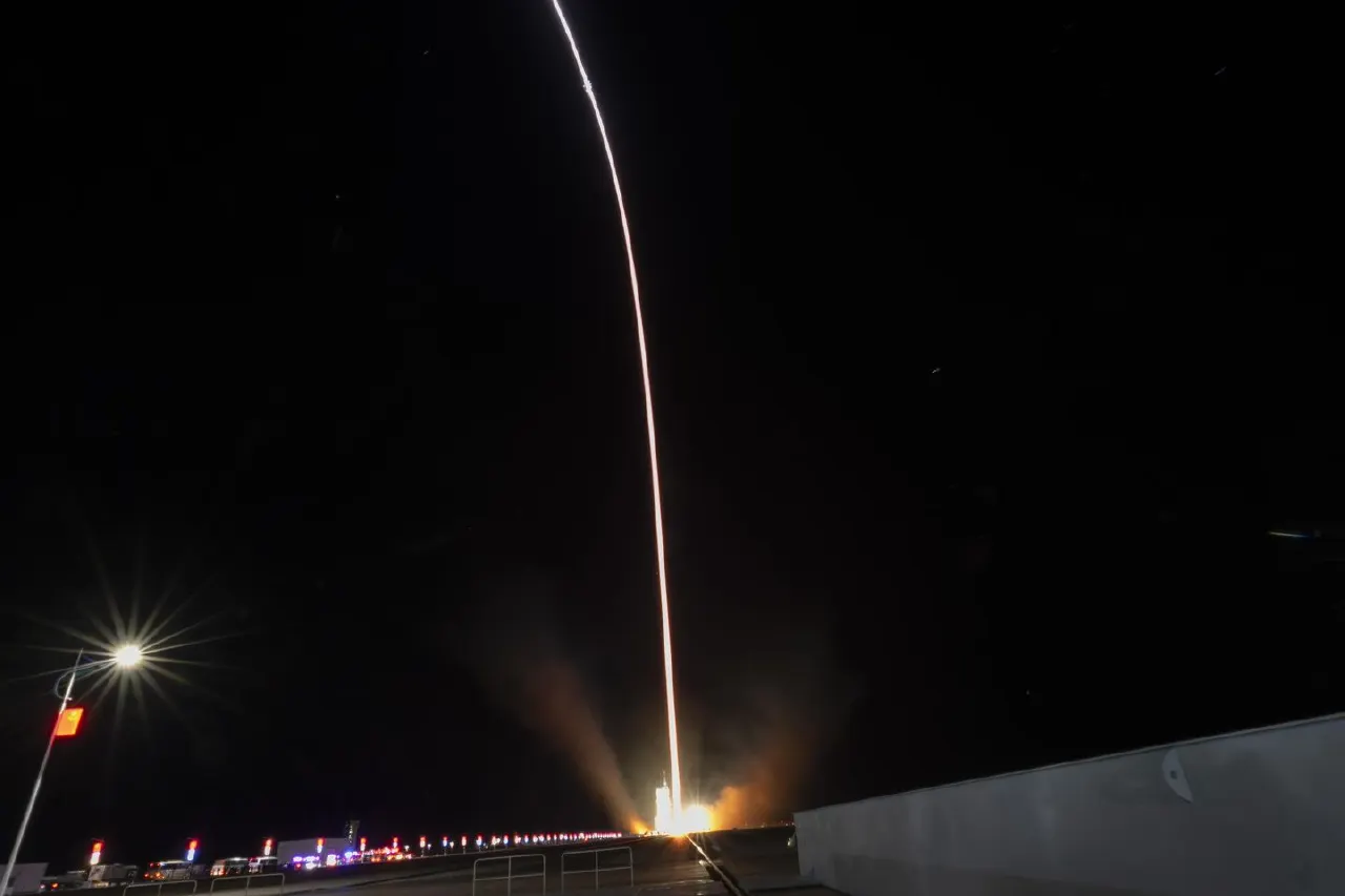 Long-exposure night photo of a rocket launching, bright exhaust trail rising into dark sky.