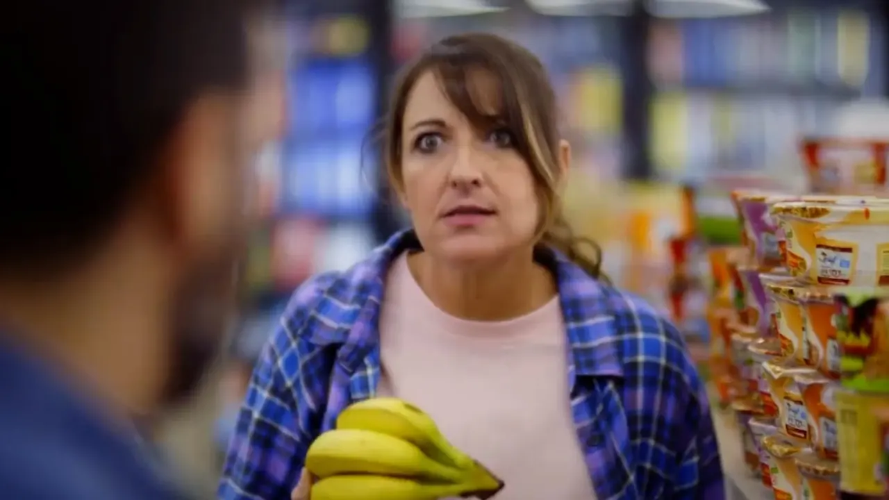 Woman with wide eyes holding bananas in a grocery aisle, a man in foreground.