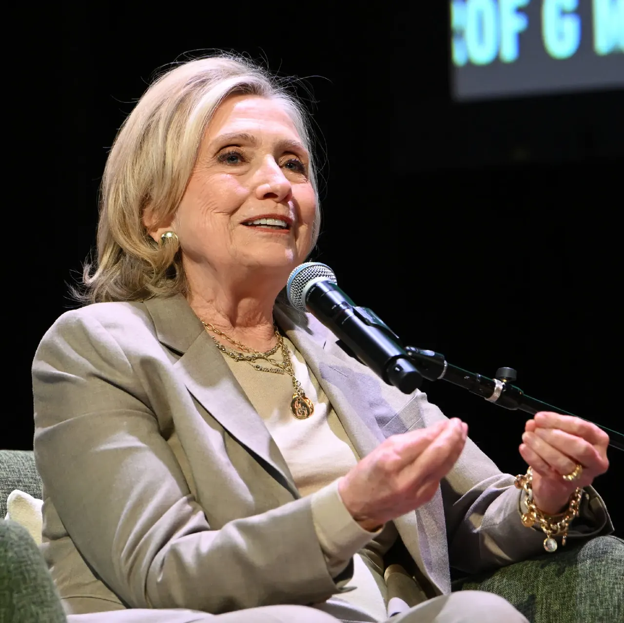 Woman in beige blazer speaking into microphone on stage, seated, smiling, with gold jewelry.
