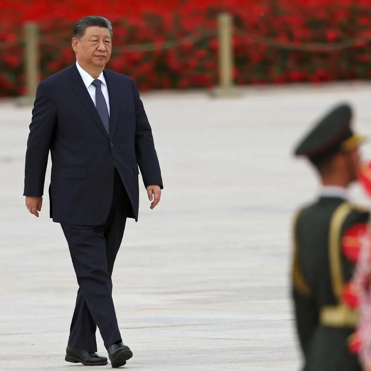 Man in dark suit walks across plaza past blurred uniformed guard, red flowers behind.