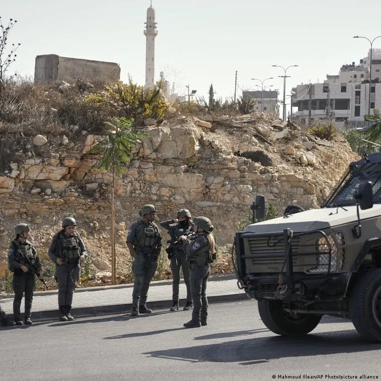 Armed soldiers in helmets and vests stand beside an armored vehicle on a street near a hillside.